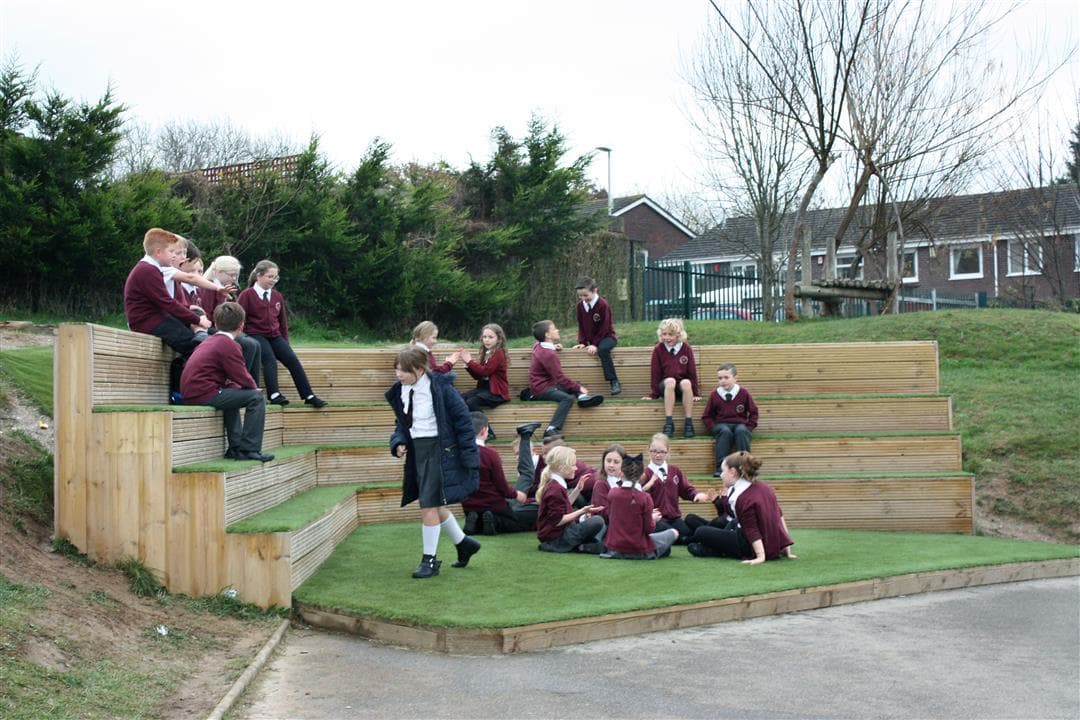 A class of children playing on their amphitheatre performance stage, artificial grass covers the top of the seats and the floor.