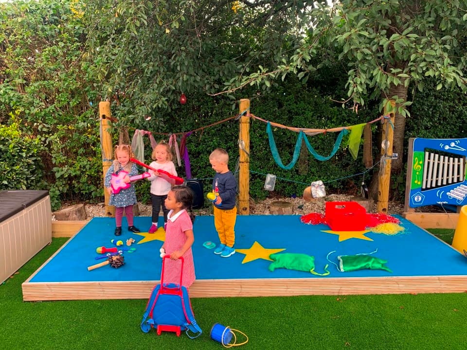 a blue performance stand with den making posts on the back, children play on the stage dancing and singing