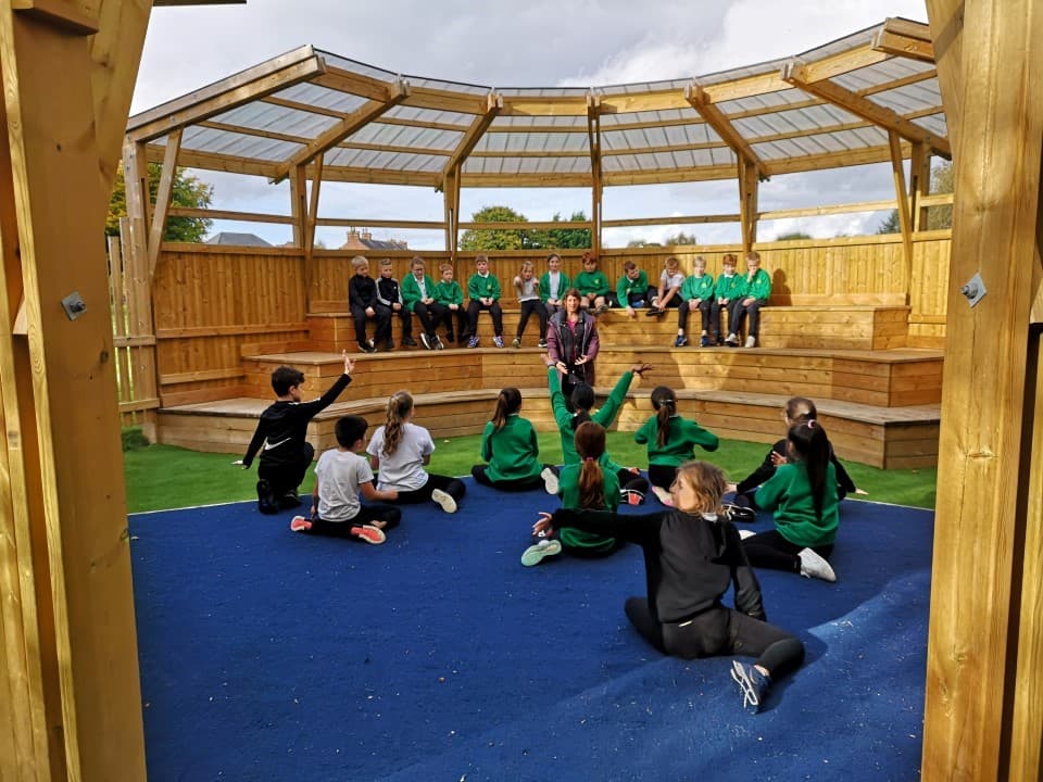 inside one of our musical ampitheatres with a performance stage and seating, children sit on the benches and watch their peers as they sit on the performance stage talking to them