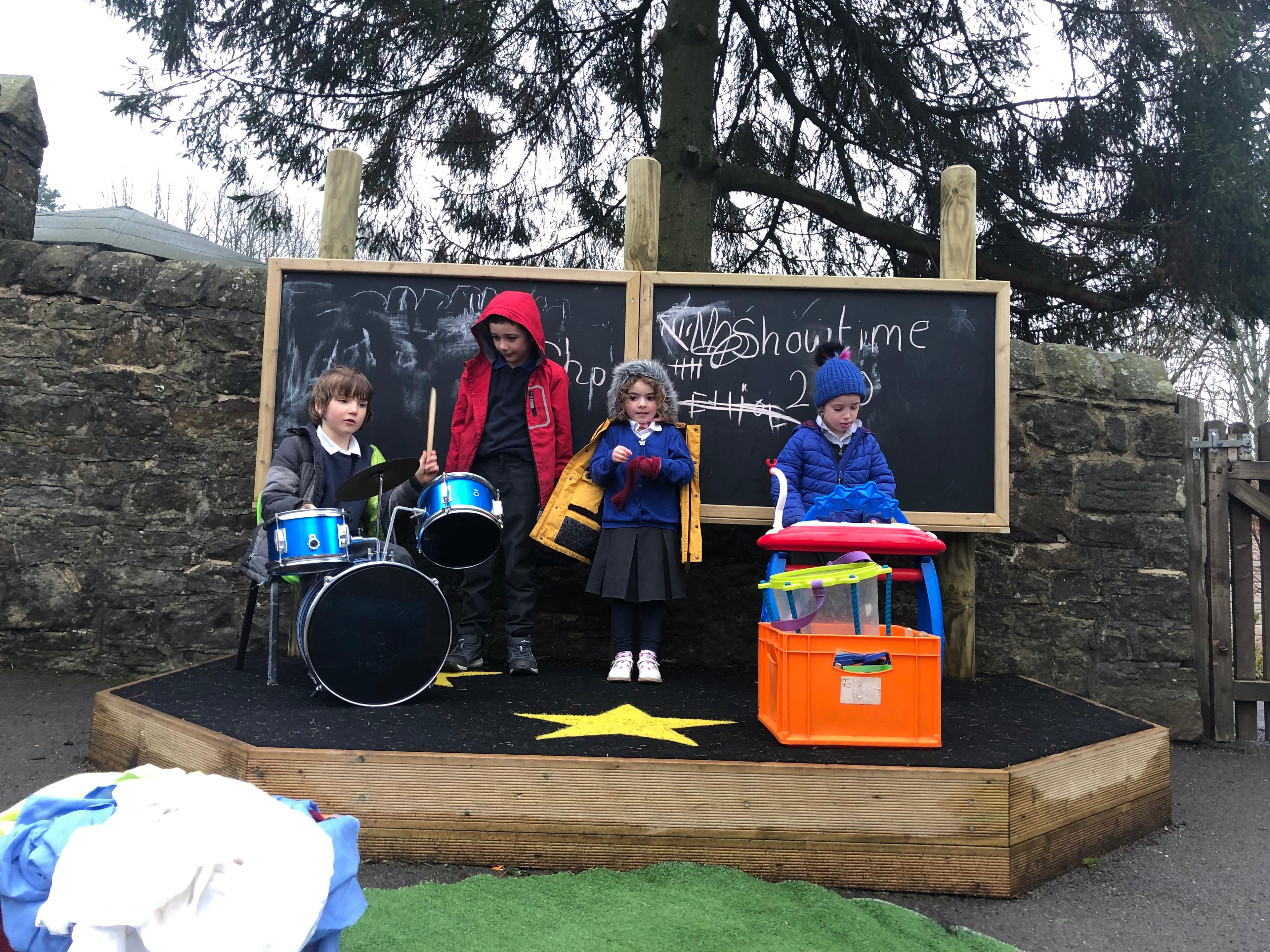 4 children playing on their new performance stage, one young boy is playing on the drums and one young boy is playing the piano.