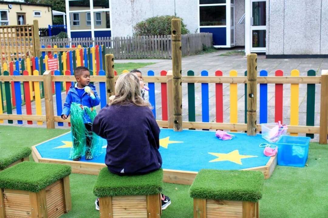 a teacher sits on artificial grass-topped seats and watches a child in a winter coat perform on the blue performance stage with rainbow fencing behind