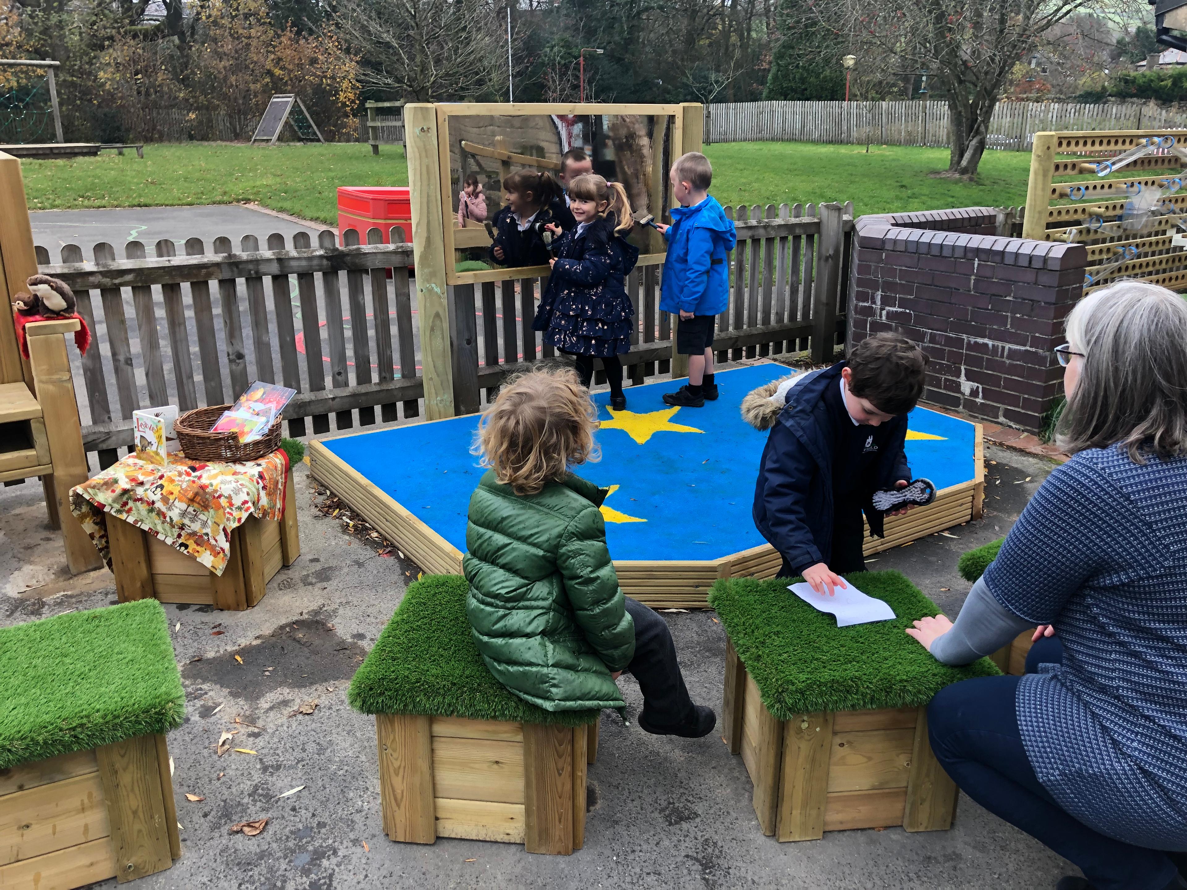2 young children playing on their new performance stage drawing on the mirror in the background. 2 children and one adult are sat on the grass top seats in front of the stage.