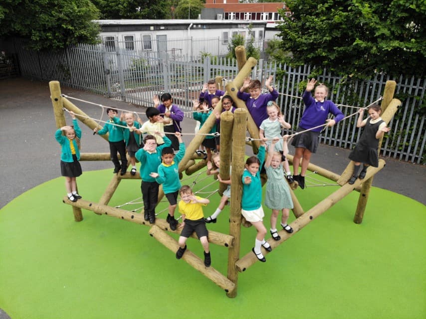 A climbing frame that has been installed on top of green wetpour on a playground. A group of children are playing on the climbing frame as they look at the camera, smiling.