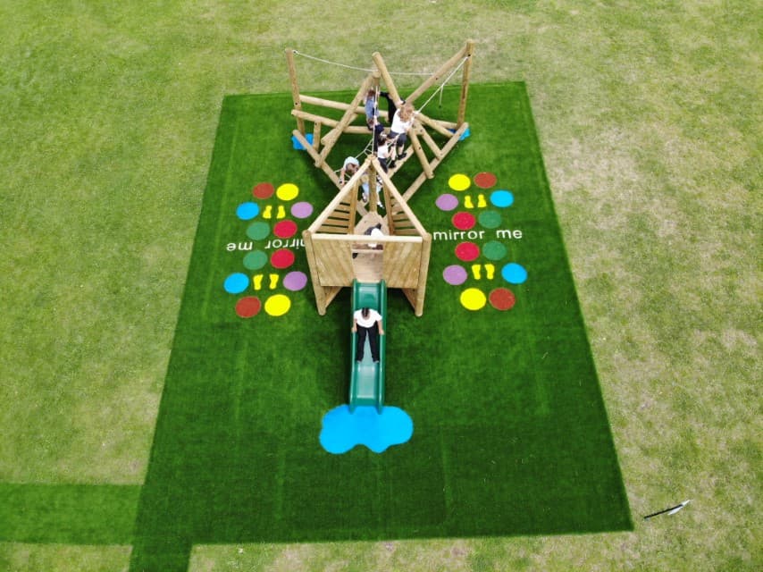 A birds-eye photo showing a climbing frame and a play tower being installed on the same patch of artificial grass. Both of these pieces of play equipment are installed right next to each other. A group of children are playing on the equipment.