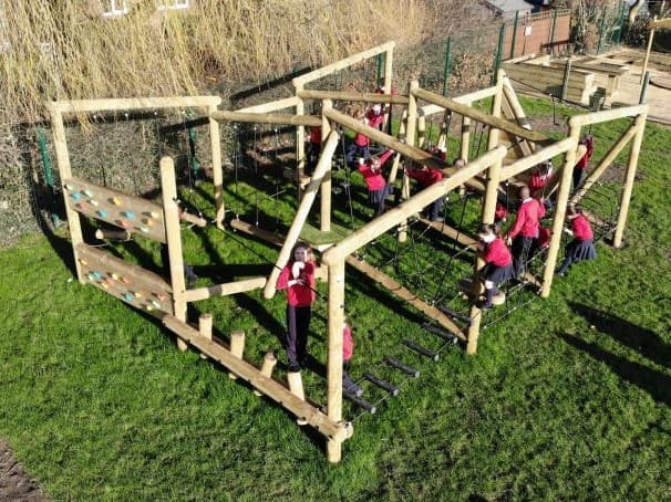 A Forest Circuit has been installed on natural grass, with a group of children playing on it. Trees can be seen in the background, with a group of planters being seen in the far distance.