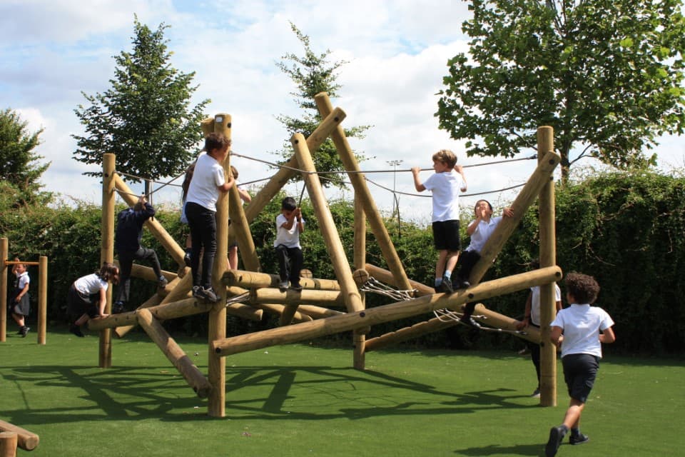 A climbing frame has been installed on an artificial grass surface. A group of children are climbing all over the equipment. Trees and bushes can be seen in the background.
