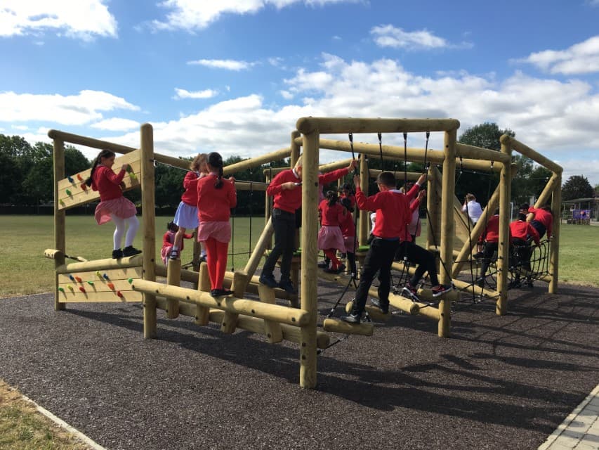 The Grizedale Forest Circuit installed on a black wetpour sub base. The circuit is quite big, with a huge group of children playing on the equipment. The Grizedale Forest Circuit is surrounded by trees and natural grass.