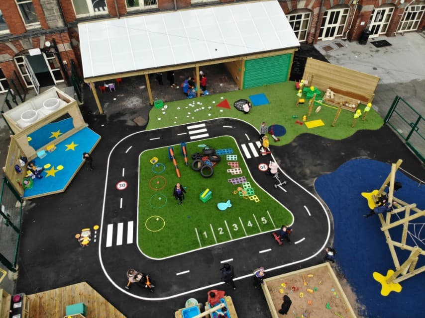A variety of thermoplastic playground markings can be seen all around a playground. The most prominent one is a road marking, that goes in a circle on black tarmac. Artificial grass can be seen on the playground too.