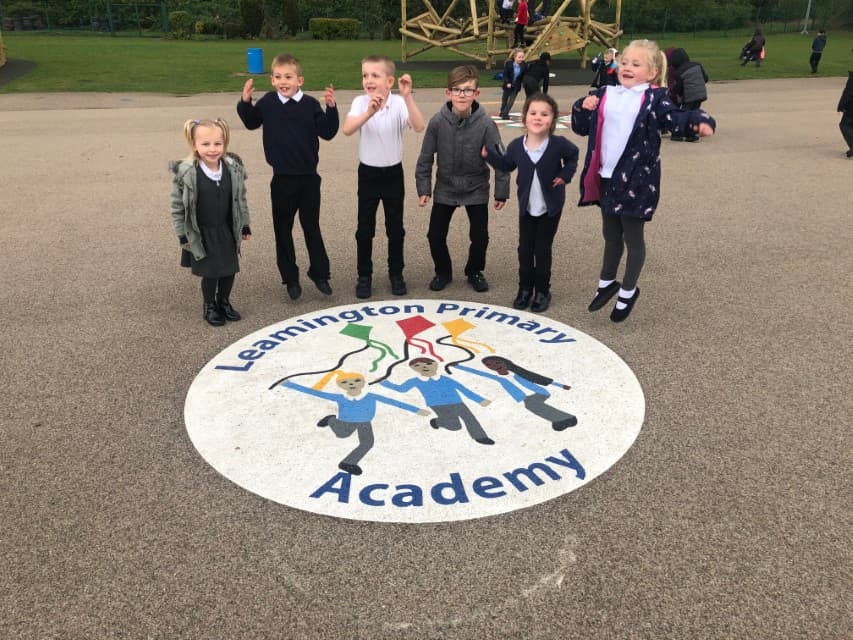 A bespoke playground marking that is a white circle, with markings of children holding kites in the middle. The text "Leamington Primary Academy" can be seen going around the edge of the circle. A group of children are stood above the graphic, smiling and waving at the camera.