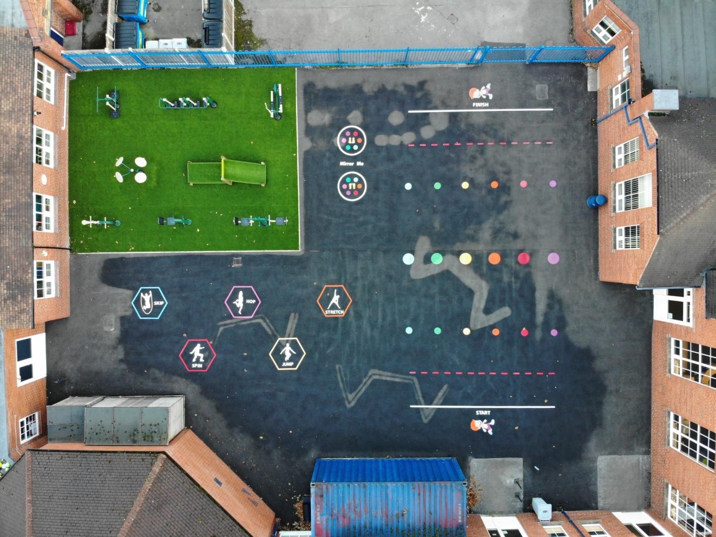 A birds-eye shot of a playground that shows a variety of different playground markings. In the top left corner, an artificial grass surface can be seen. The thermoplastic markings have a variety of different colours.