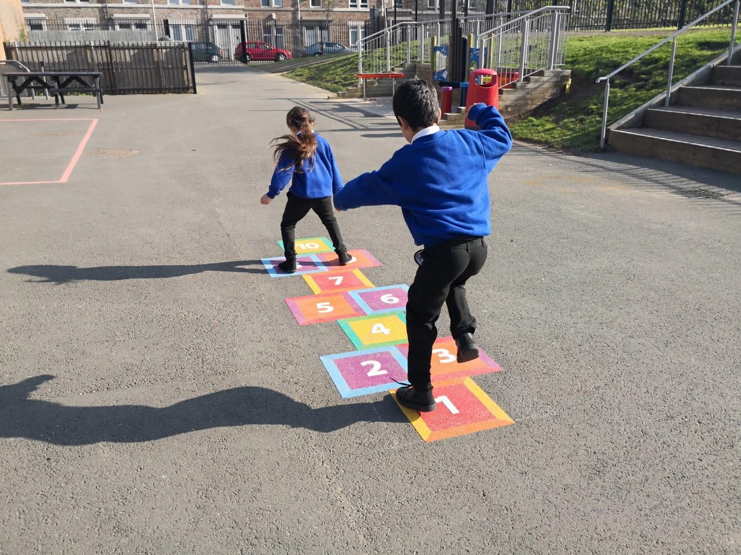 2 primary school children are playing on a thermoplastic playground marking of a hopscotch graphic. The thermoplastic graphic has been installed on a tarmac surface,