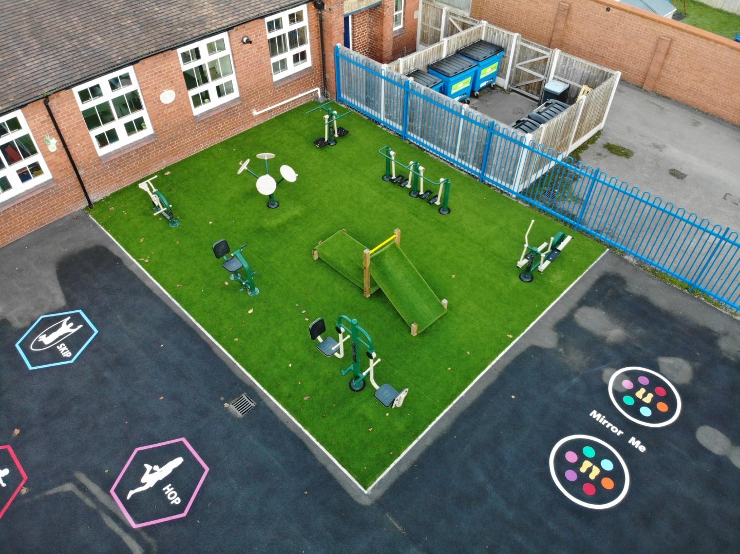 Astroturf installed in the corner of a playground, with gym equipment being placed on the surface, as well as two other pieces of wooden play equipment. Surrounding the artificial grass is a tarmac playground, with thermoplastic play markings.