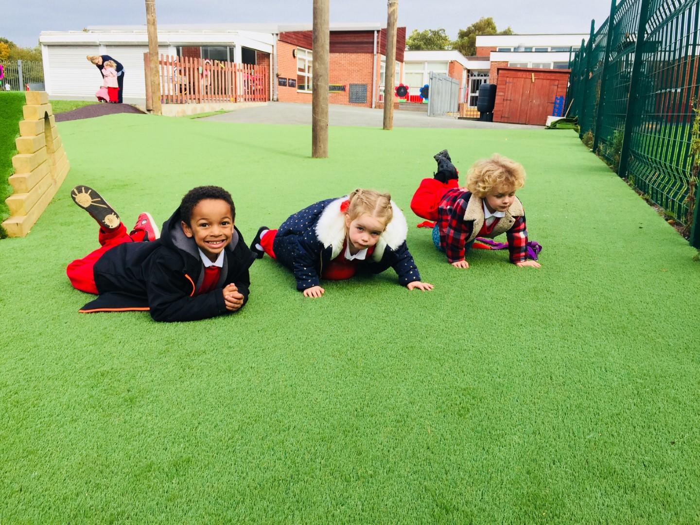 3 children are laid down on an artificial grass surface, looking into the camera. The children are smiling as play equipment can be seen in the background too.