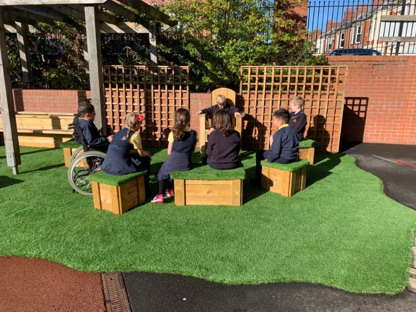 A variety of wooden blocks with artificial grass tops have been placed in a circle, with a giant wooden chair being placed at the end of the circle. All the children are looking at the person in the wooden chair. A child in a wheelchair is sat next to another child who is sat on a wooden block.