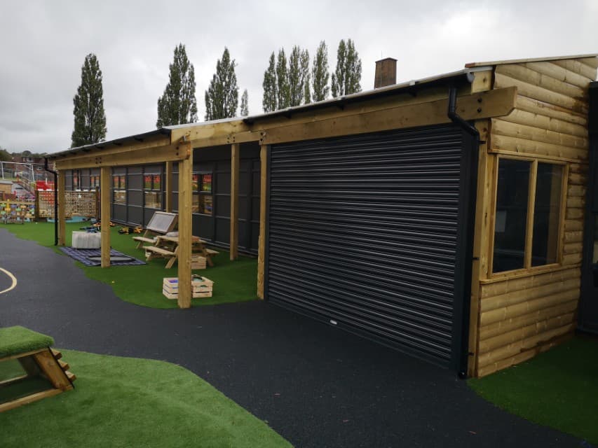 A timber canopy is built on top of a surface of wetpour and artificial grass. The canopy has a metal shutter to protect outdoor equipment. It also has windows and walls, allowing for classes to use the space for learning.