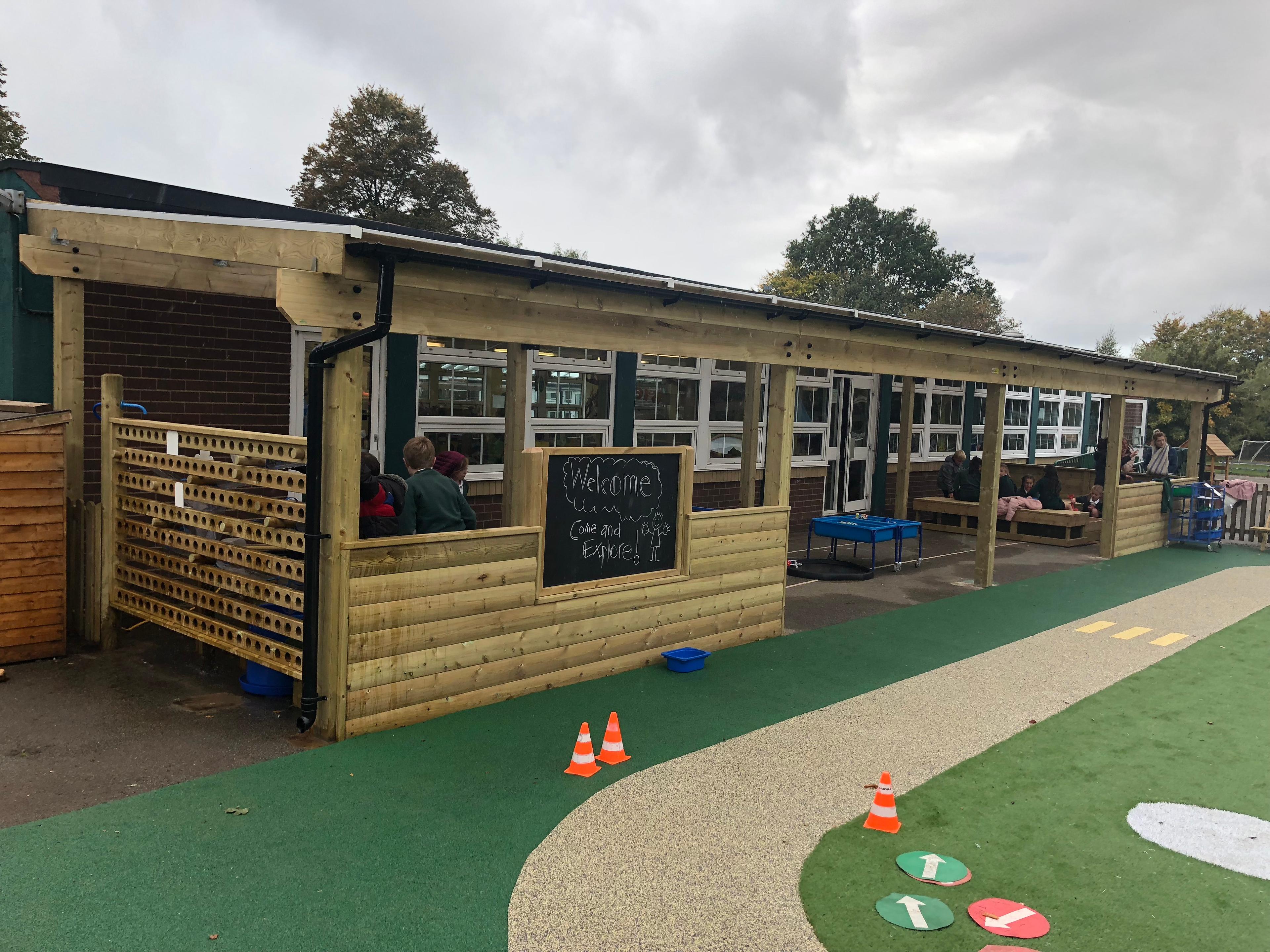 A wooden canopy has been installed on a wetpour surface. The canopy has been connected to a school, with a variety of outdoor play equipment stored underneath it. The canopy is huge, with a chalkboard installed on one of the walls.