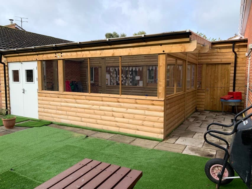 A wooden canopy installed on an artificial grass surface. It has double white doors on the entrance, with a row of windows going around the whole structure. A variety of outdoor equipment can be seen scattered around the play area that lies in front of the building.
