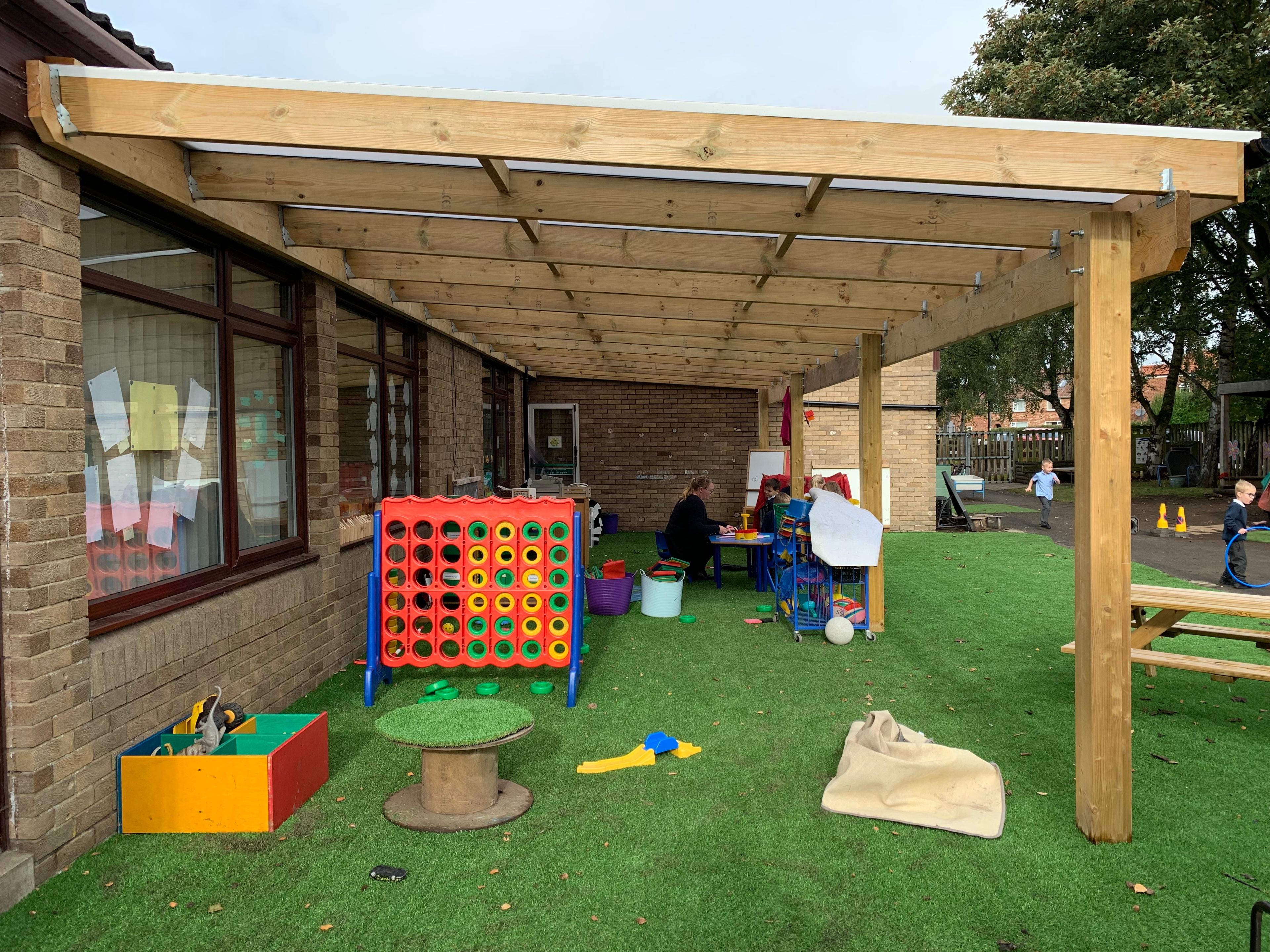 A timber canopy is attached to a school building. A wide variety of outdoor toys and learning equipment can be seen underneath the canopy. The surface underneath the canopy is artificial grass.