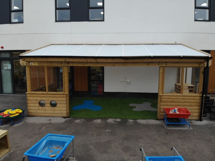 A wooden canopy is installed on top of a concrete playground, with an artificial grass surfacing installed inside the structure. The canopy is connected to the school and has a white roof, with windows and walls, and has a big entrance at the front.