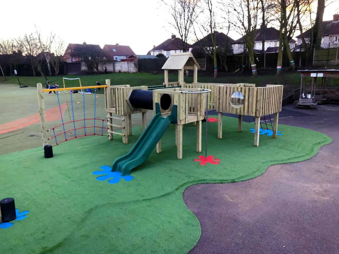 A play tower that has been installed on a layer of Saferturf with a variety of Saferturf graphics scattered around the area. A climbing net and slide can be seen from the front of the wooden structure.