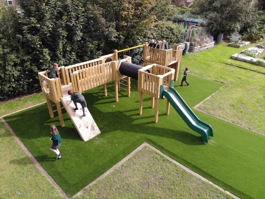 A wooden play tower that has been installed on an artificial grass playground. A group of children are playing on the equipment and engaging with different areas of the play equipment.