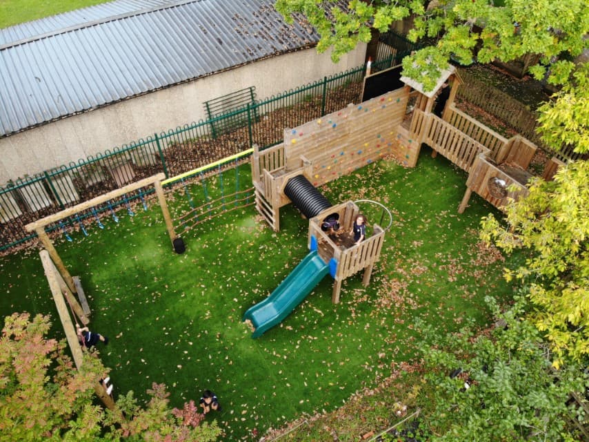 A play tower with a trim trail attached to it. The equipment has been installed on a natural grass surface. Some children can be seen playing on the equipment. A school building can be seen behind the equipment.