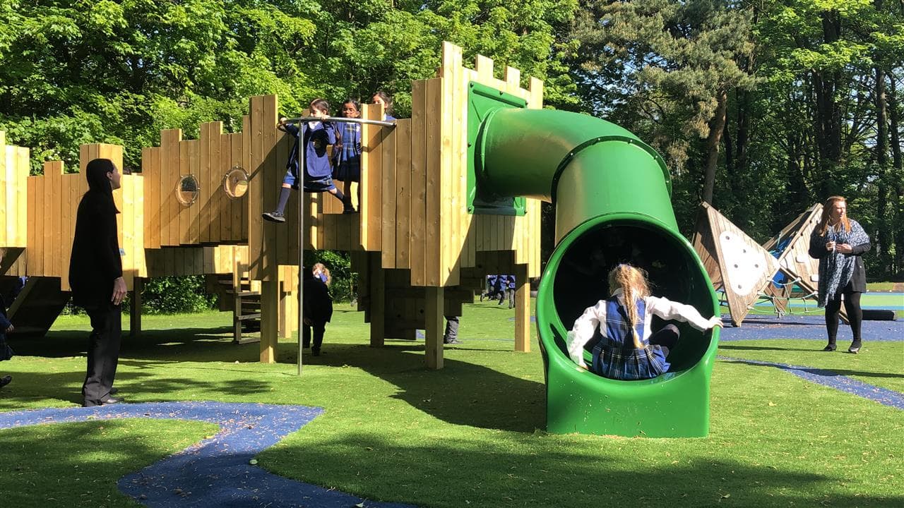 3 children playing on the tree house, one young boy is sliding down the slide, the two other children are sliding down the pole on the side. An adult is accompanying them. 