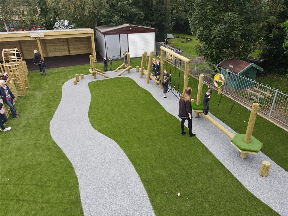 A birds eye view of a class of children playing on their trim trail, 1 teacher is stood to the side accompanying the children. Artificial grass covers the play area, with a wetpour road design placed underneath the Trim Trail itself.