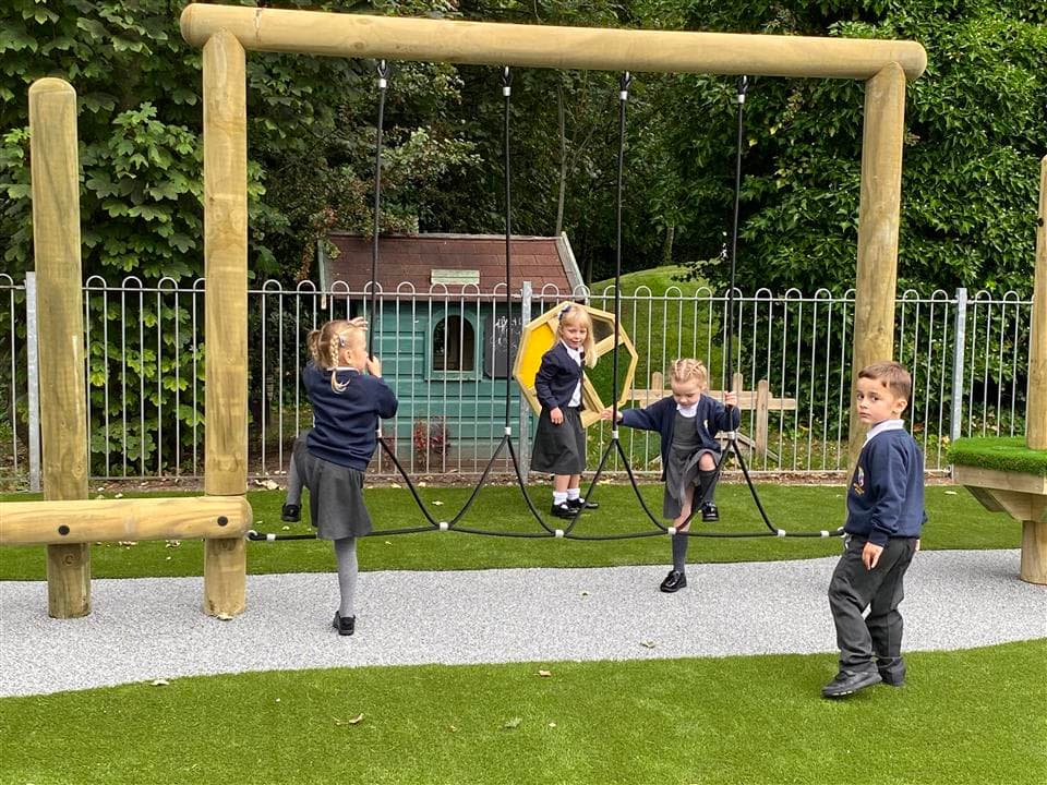 3 children playing on their trim trail, 1 young boy is playing on the side. A layer of artificial grass can be seen on both sides of the Trim Trail, as a thin strip of grey wetpour is laid underneath the trim trail.