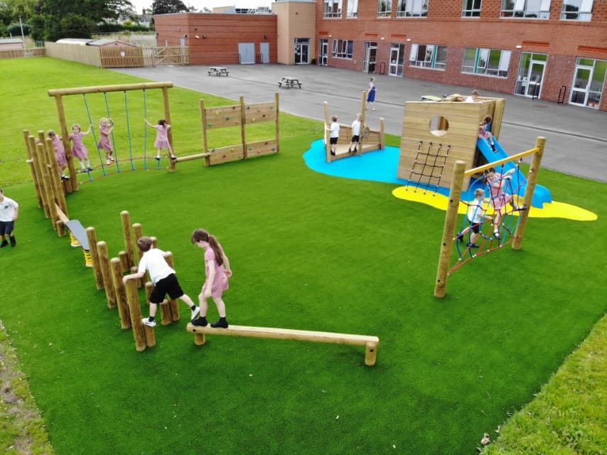 A class of children playing on different pieces of equipment on the trim trail, the trim trail is in a circle pattern and installed on top of a layer of artificial grass.