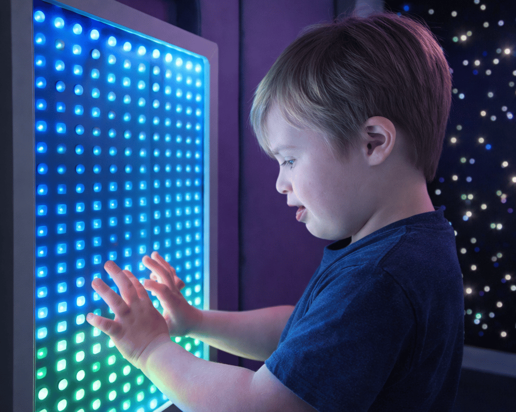 A child with Down Syndrome is touching the LED infinity mirror, as he stares into it, smiling.