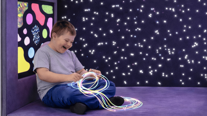 A SEN child is holding a string of lights as he laughs, leaning against the sensory wall.