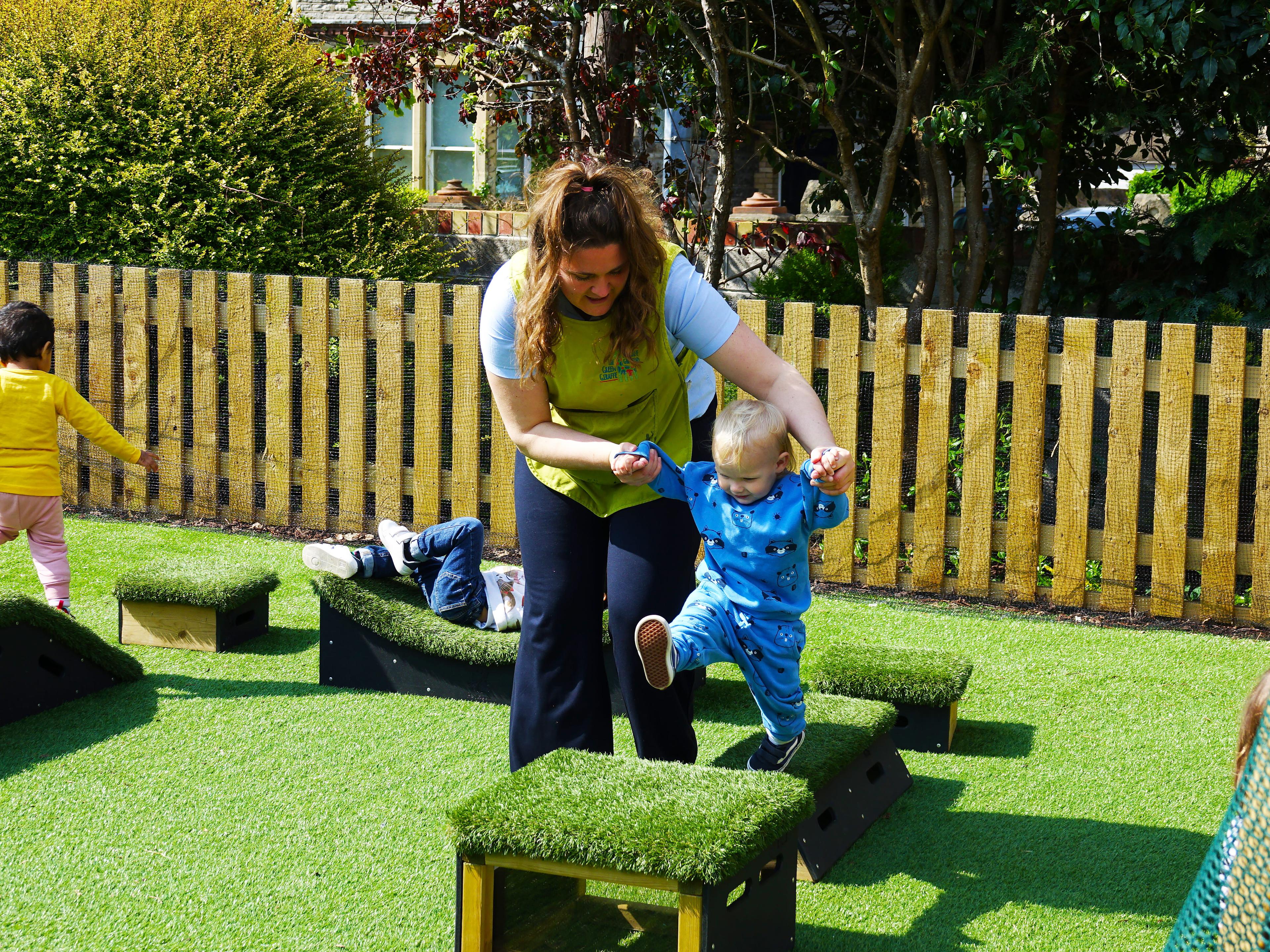A photo of a preschool teacher helping a young toddler to jump across get set go blocks.