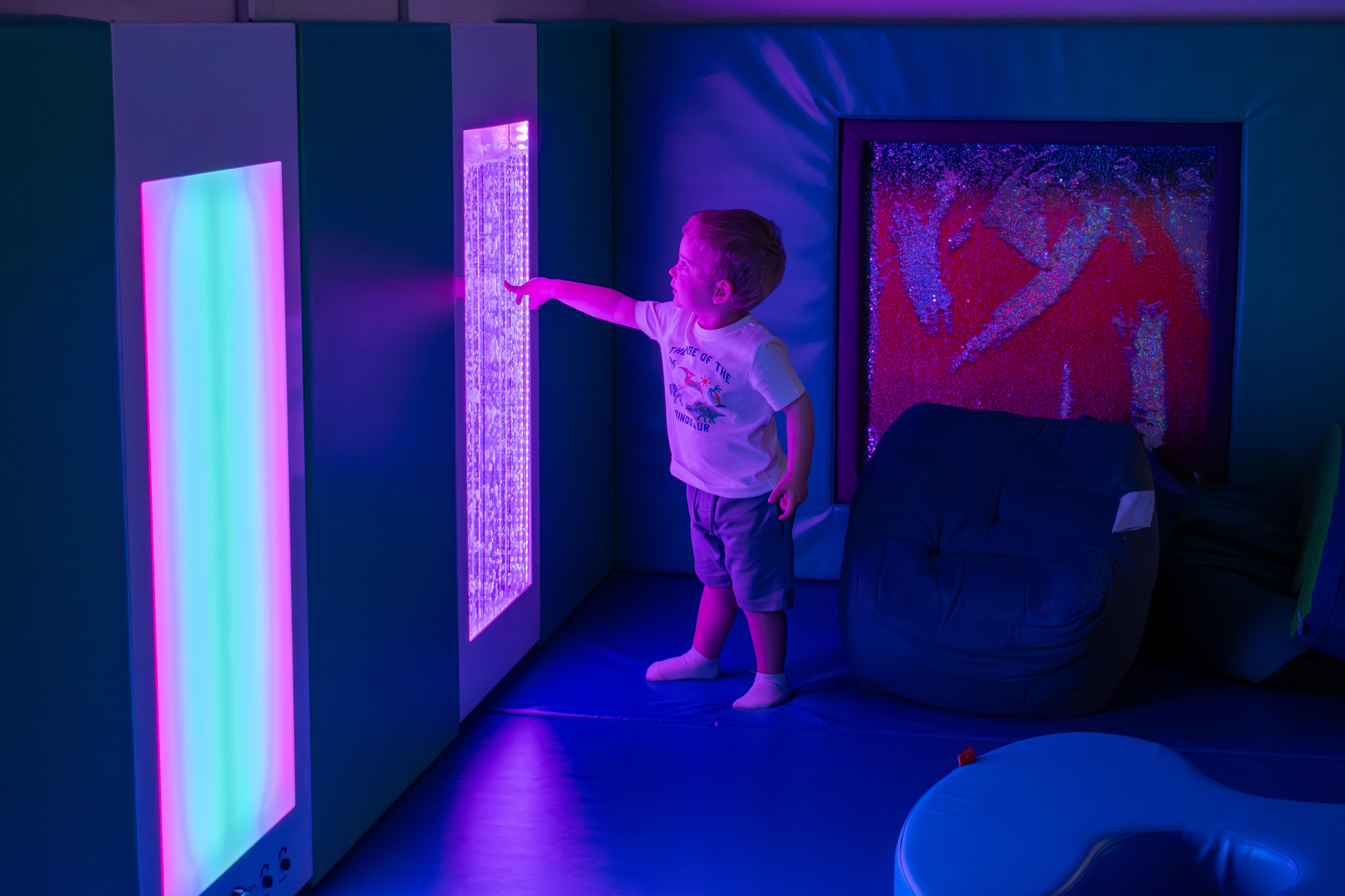 A toddler pointing at a hurricane panel in a purple lit sensory room.
