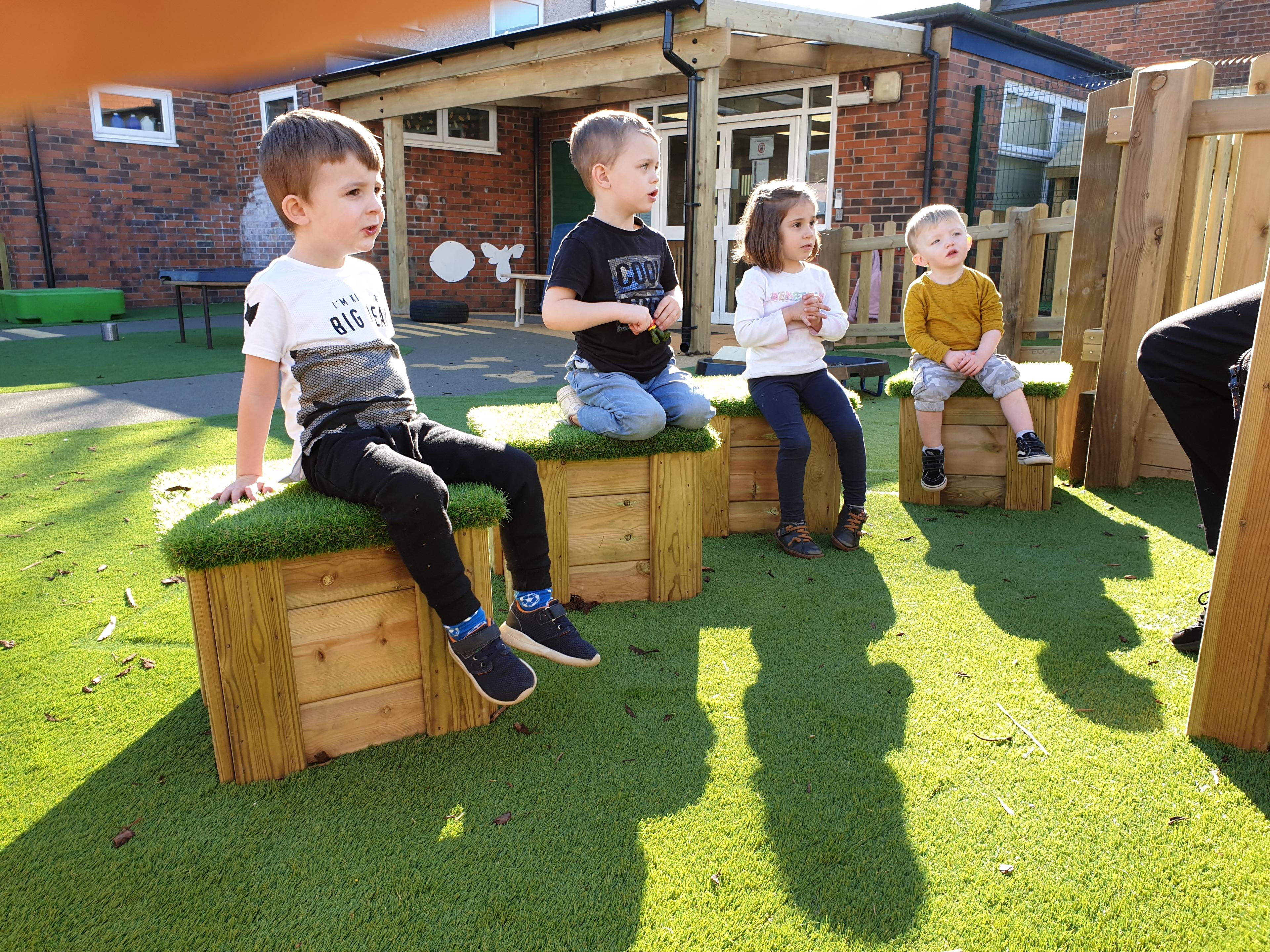 A photo of a group of nursery children sat on artificial grass-topped wooden seats.
