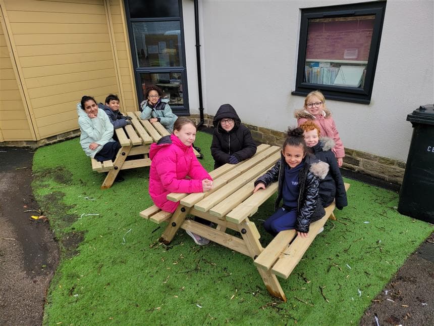 A pair of picnic tables on artificial grass with children sat smiling on them.