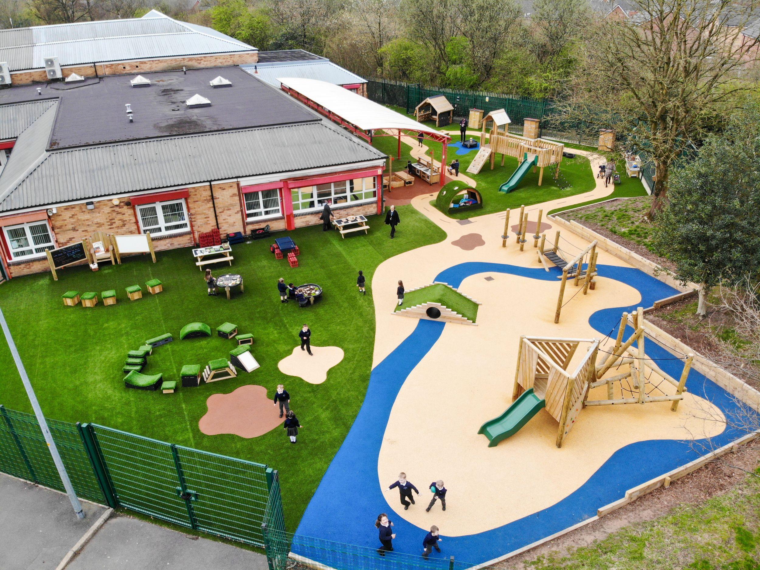 KS1 playground with multiple freestanding products including Get Set Go Blocks on artificial grass, plus two installed play towers and trim trail. Blue and buff bespoke surfacing creates a winding "river" path through the active zones.