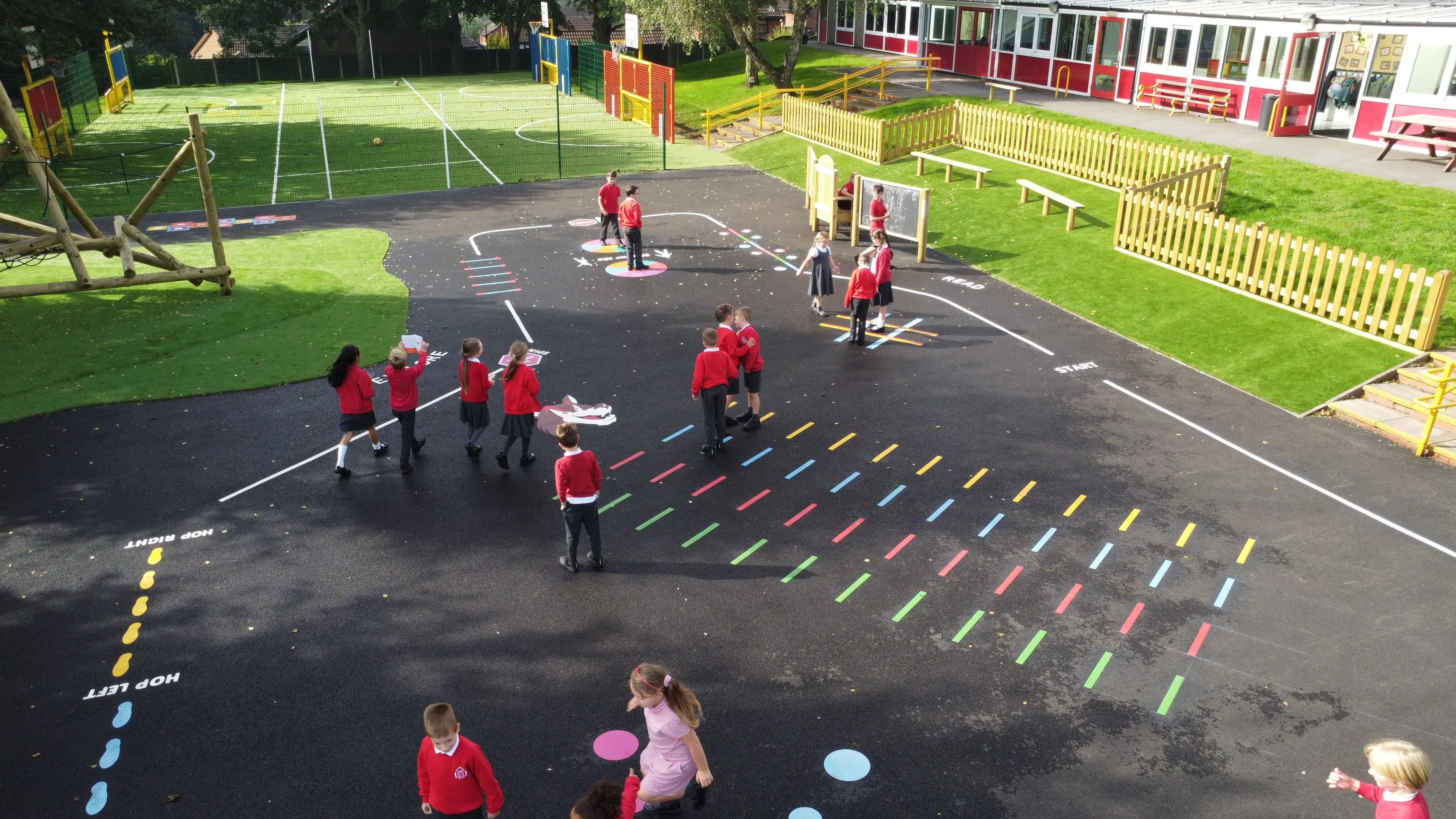 A playground with a variety of different playground markings installed on it. A line goes around the playground as a trail. A group of children are playing on the playground markings.