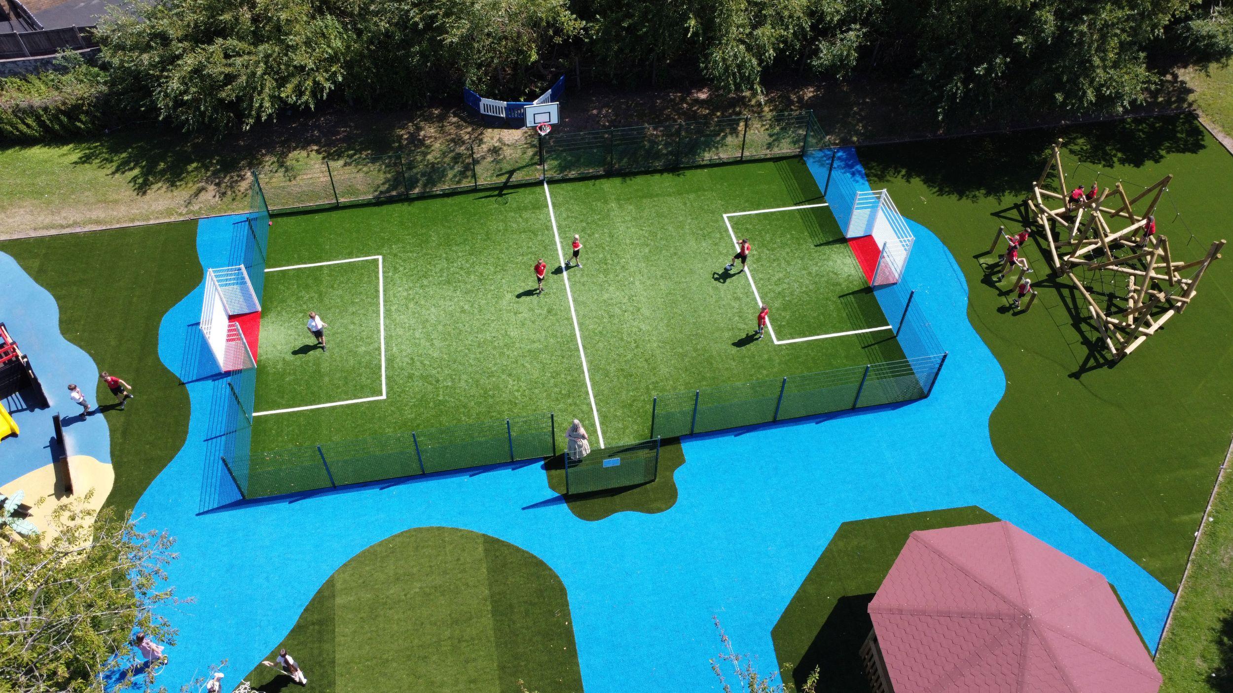 Aerial view of a KS2 MUGA on artificial grass, surrounded by blue Wetpour. A large wooden Helm Crag Climber and an Octagonal Gazebo sit in separate zones within the same vibrant playground design.