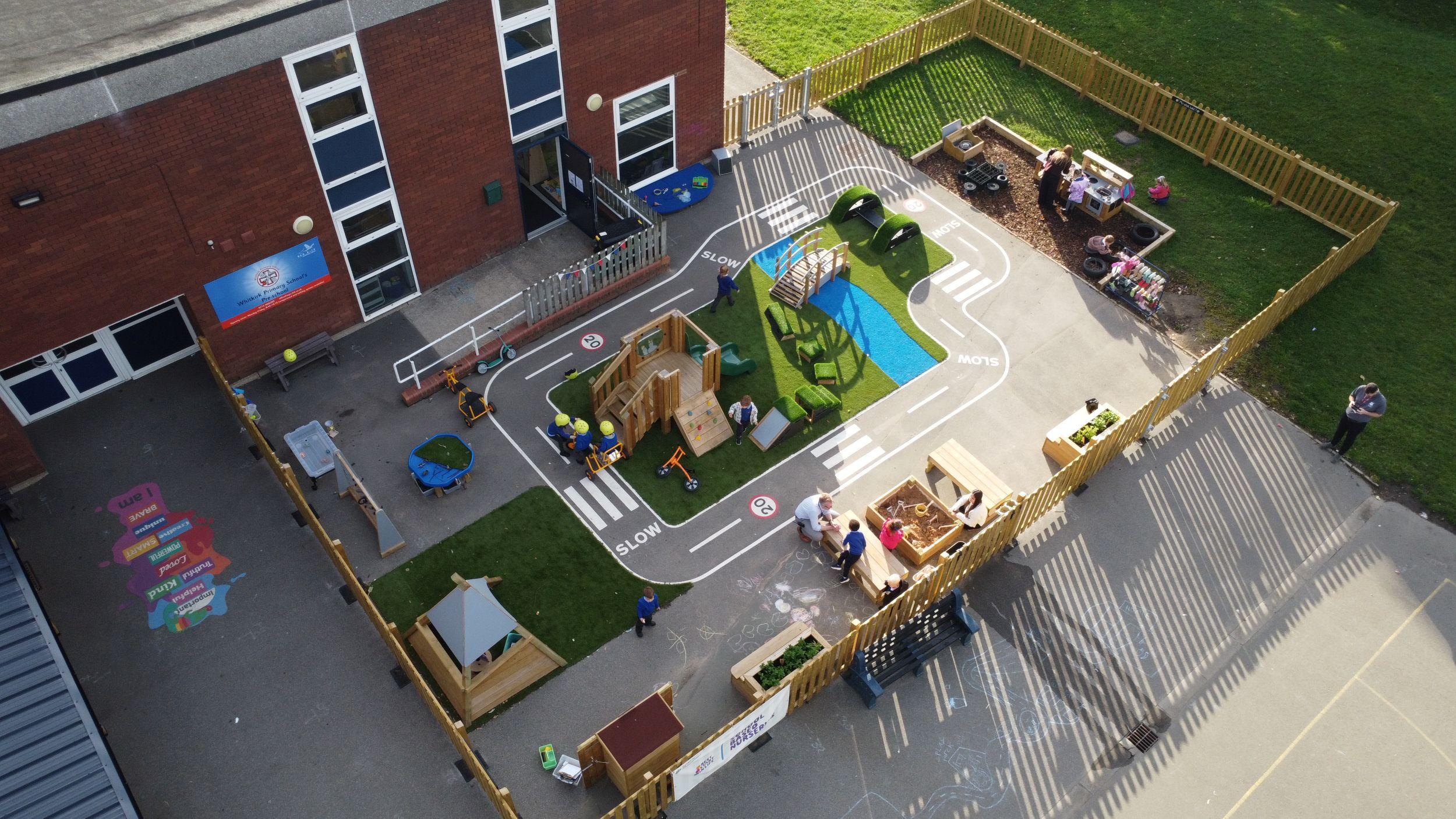 Nursery scheme with a Restormel Play Tower and Play Bridge with Pentagon Play's "Rockies" on artificial grass. A track connects the zones, with separate Mud Kitchen and Wigwam areas on the playground's edge.