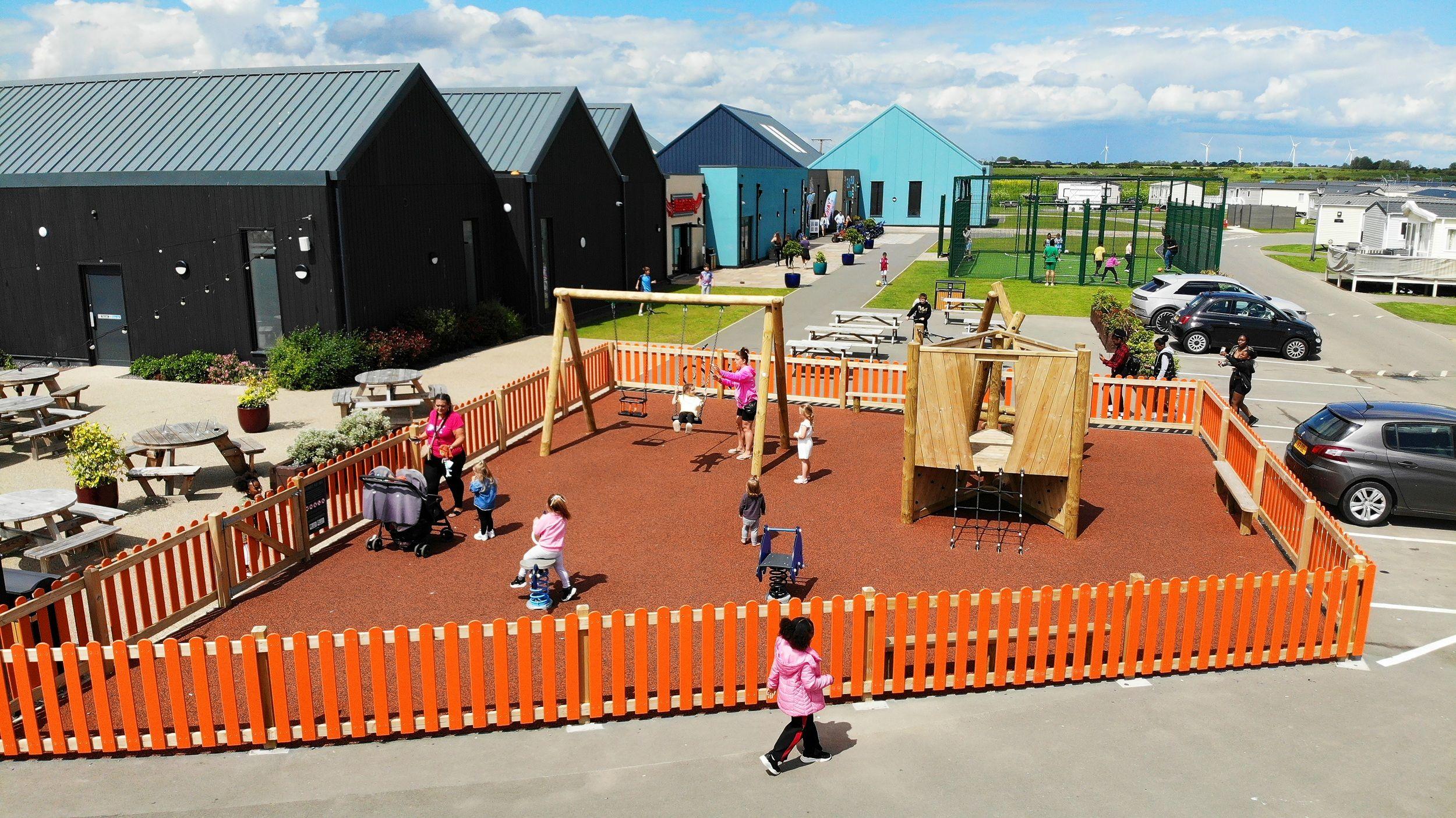 Children play on a wooden swing set and climber on red wetpour, enclosed by an orange fence. Modern buildings and a MUGA sit in the background of this active holiday park scene.